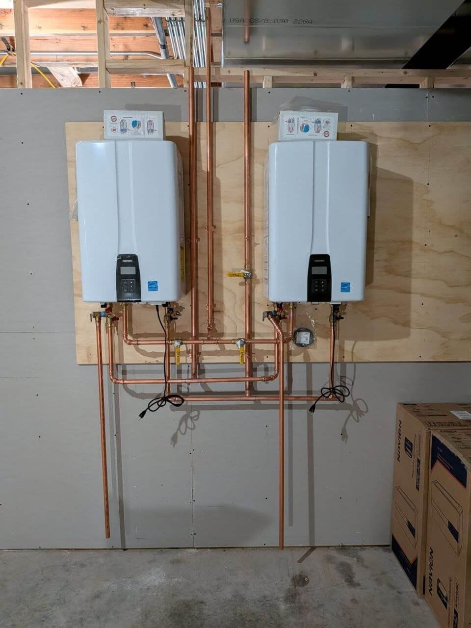 Two wall-mounted tankless water heaters with copper piping on a plywood backboard